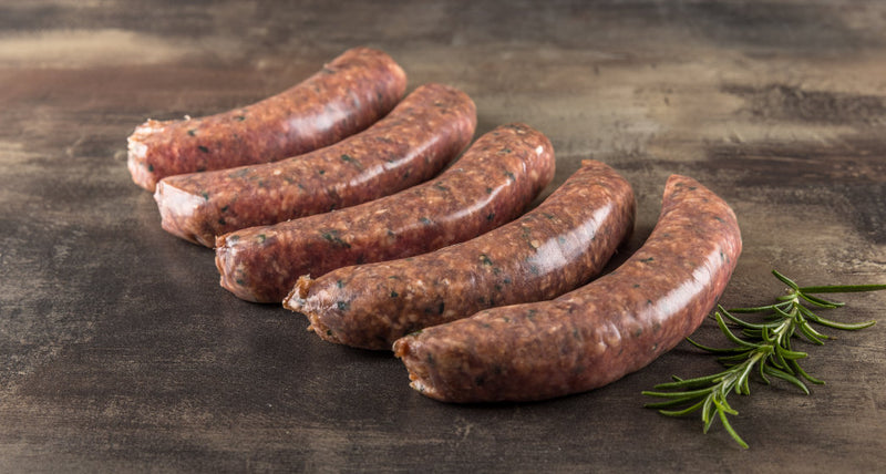 Fünf rohe Pure Wagyu Salsiccia im Naturdarm auf dunklem Stein, daneben ein Rosmarinzweig