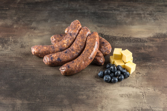 Gestapelte rohe Blueberry BBQ Salsiccia auf dunklem Stein, daneben Gouda-Würfel und Blaubeeren
